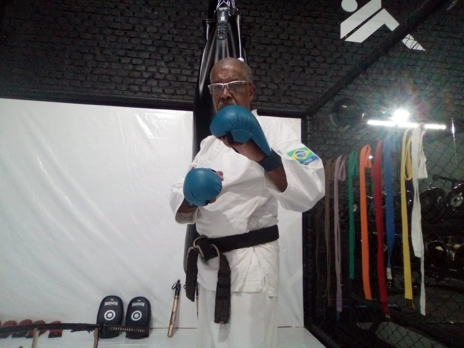 O professor do Delta CT, de kimono e faixa preta, em guarda diante do saco de pancada, com as faixas coloridas na parede.